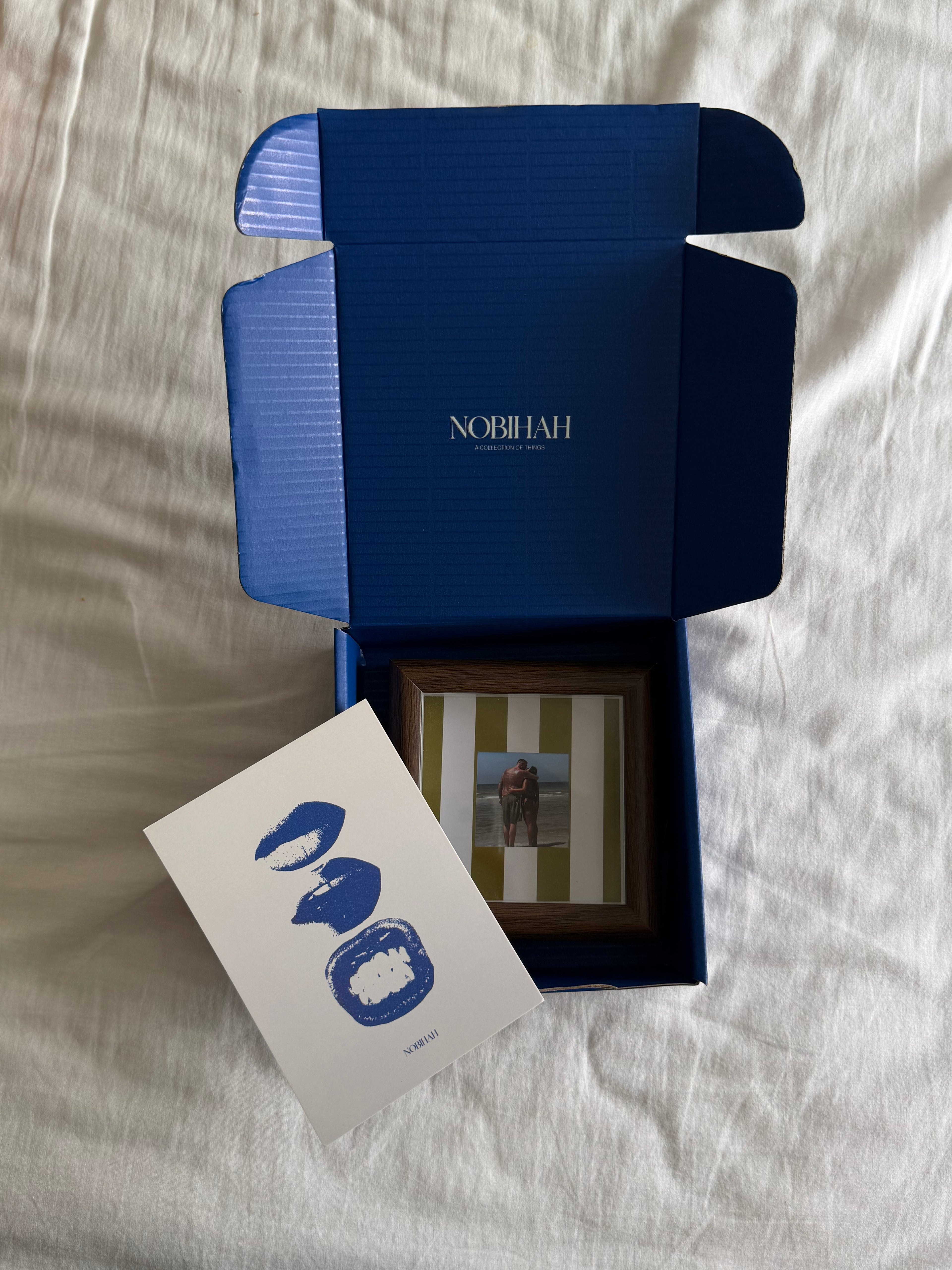 An open dark blue gift box on a white fabric surface, containing a small framed photo and a white card with blue abstract shapes. The inside lid shows the brand name NOBIHAH.