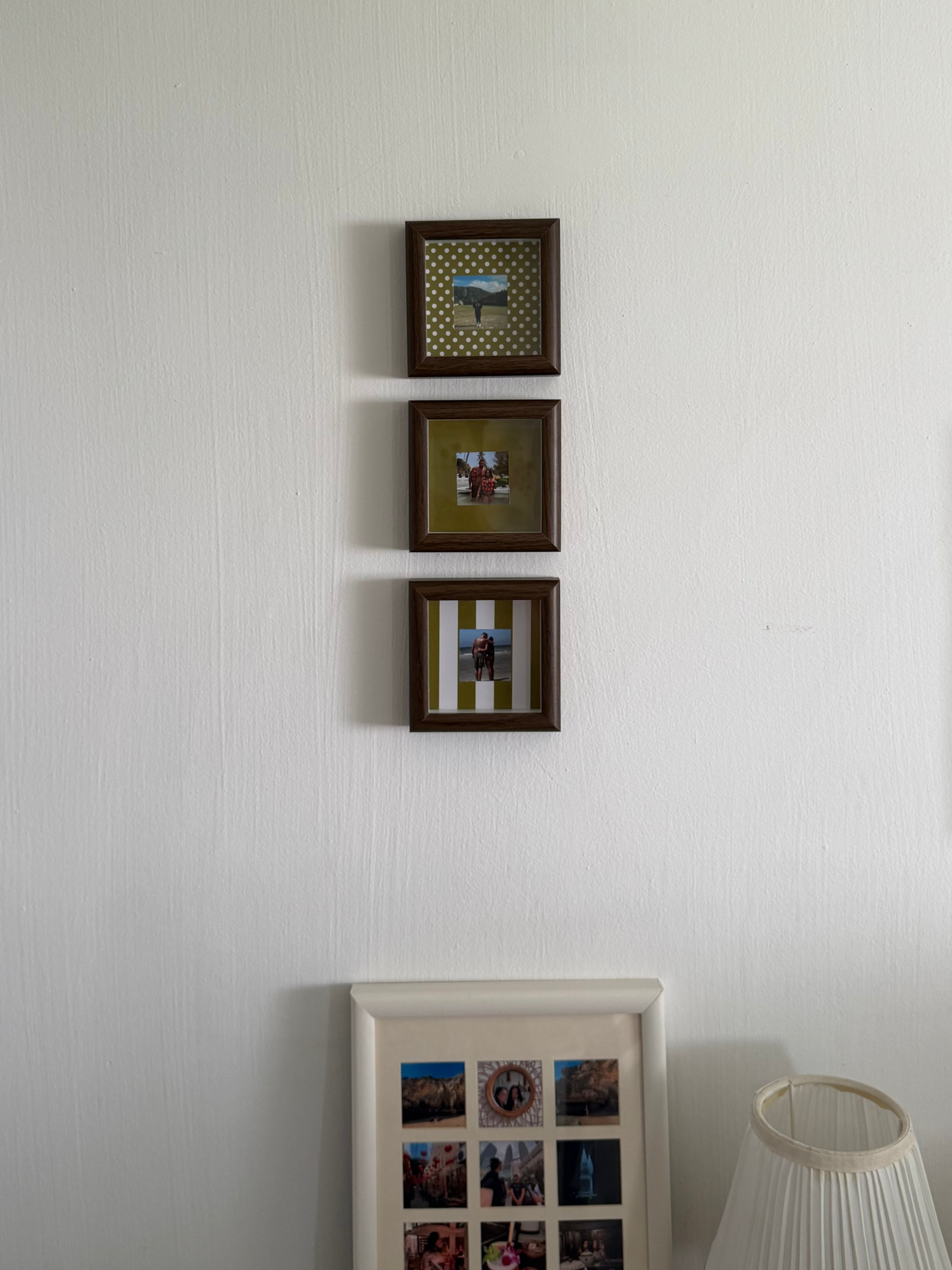 Three small square framed pictures with dark brown frames hanging vertically on a white textured wall, with a larger white frame and a pleated lamp visible below.